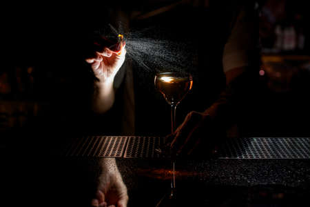 Professional male bartender spraying on the brown alcoholic drink with a orange zest juice in the darkの写真素材