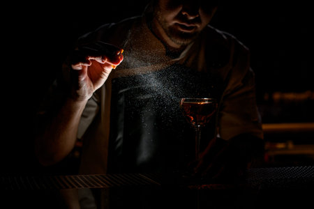 Professional bartender spraying on the brown alcoholic drink with a orange zest juice in the dark