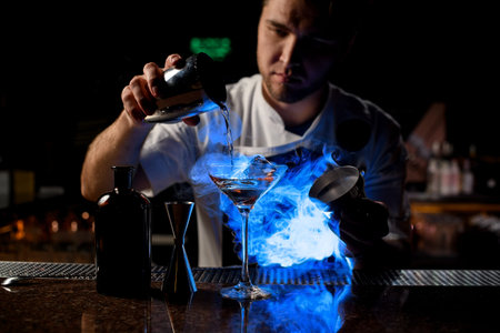 Professional bartender pouring a brown alcoholic drink from the steel shaker to the glass under blue...