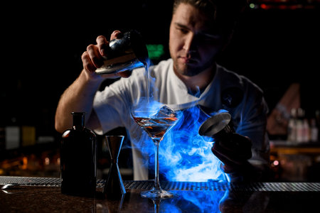 Bartender pouring a brown alcoholic drink from the steel shaker to the glass under blue light