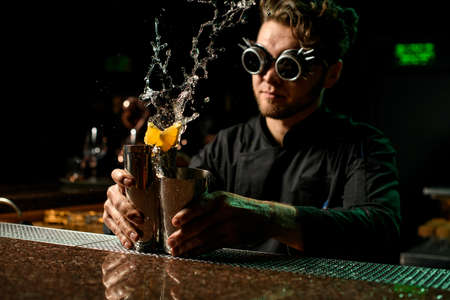 Bartender holding in hands a steel shakers splashing the cocktail with orange pieces in it on the bar counterの写真素材