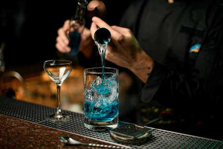 Professional male bartender pouring a blue alcoholic drink from the jigger to a measuring cup holding a glass bottleの写真素材