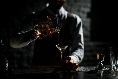 Professional male bartender pouring a golden alcoholic drink from the measuring cup with straner to...