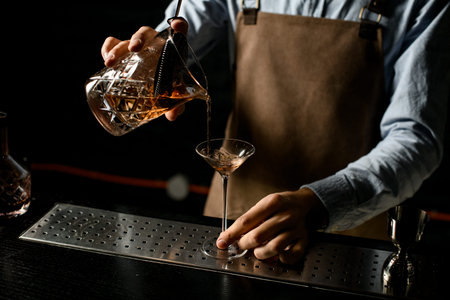 Male bartender pouring a brown alcoholic drink from the measuring cup with straner to a martini...