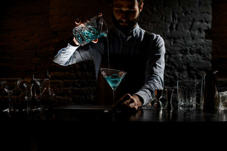 Professional bartender pouring a blue alcoholic drink from the measuring cup with strainer to a martini glassの写真素材