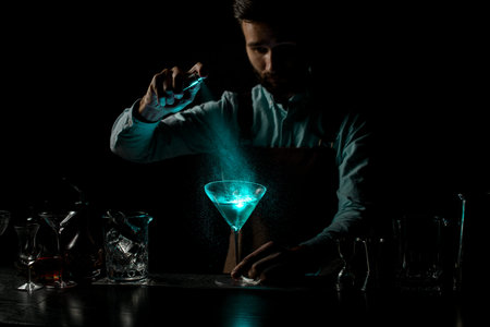 Male bartender spraying with a bitter on a blue alcoholic cocktail in a martini glass decorated with...