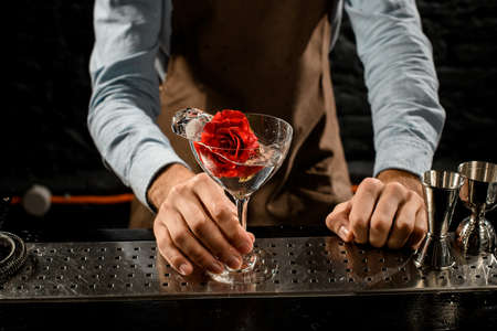 Professional male bartender serving a martini glass with a cocktail decorated with red rose budの写真素材