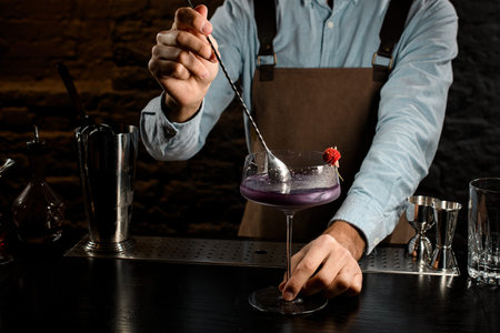Male bartender stirring a violet alcoholic drink in the big cocktail glass decorated with a red...
