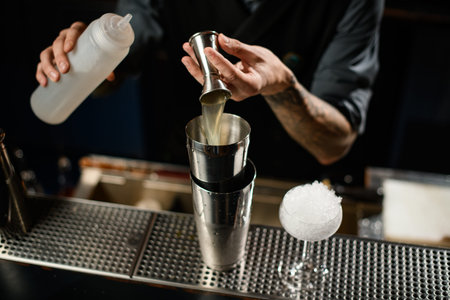 Bartender pouring a alcoholic drink from the jigger to a steel shaker holding a white plastic bottle