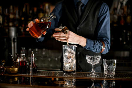 Bartender measures the ingredients for cocktail from bottle with brown alcohol drink.の写真素材