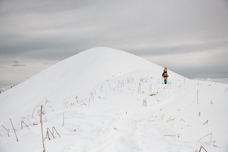 man with snowboard on his back climbs along path to snowy mountain.