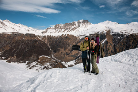 Couple of travellers with snowboards posing on snowy top of mountains