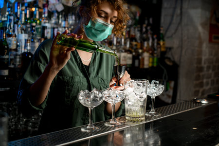 Girl bartender in medical mask pour liquid from bottle into beaker.