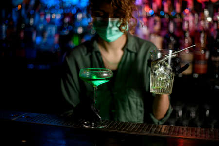 young girl at bar counter in medical mask and black gloves holds glass with green cocktail.の写真素材