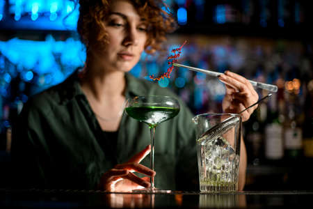 young woman at bar holds glass with cocktail and carefully decorates itの写真素材