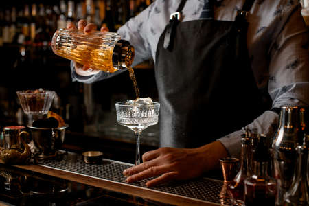 Professional barman pours finished cocktail from glassy shaker into glass with ice.の写真素材