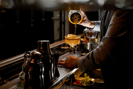 Professional barman carefully pours finished cocktail from glassy shaker into glass with ice.