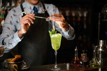 Professional bartender decorates green alcoholic drink with ice in glass by powder.