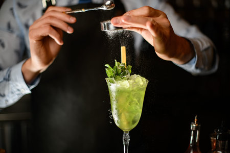 Close-up bartender decorates green alcoholic drink with ice in glass by powder.