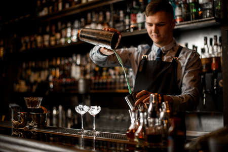 young handsome bartender pours cocktail in shaker. Two glasses with ice stand on bar.の写真素材
