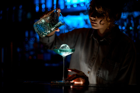 Woman at bar attentively pours cocktail into glass.