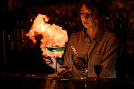 Woman at dark bar professionally makes huge fire flame over wineglass with blue alcoholic cocktail.の写真素材