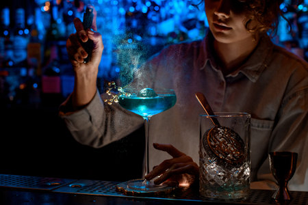 Woman bartender carefully sprinkles on glass with blue cocktail which decorated flowering plant and...