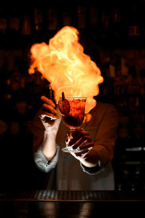 Woman at bar holds in her hand wineglass with cocktail, sprinkles on it and makes big fire over it.の写真素材