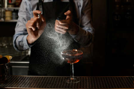 Bartender in black apron sprinkles on glass with cocktail.の写真素材