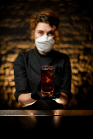 Close-up. Woman at bar in medical mask and black gloves present glass with cocktail.の写真素材