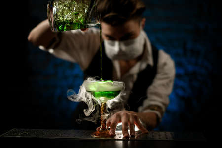bartender with white medical mask on his face pours drink of green colour into glass and attentively look at it.の写真素材
