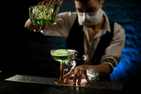 Young bartender with white medical mask on his face pours green alcoholic drink to glassの写真素材