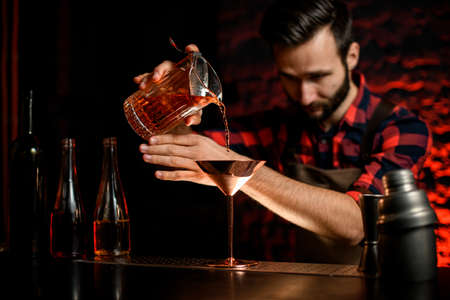 male bartender in red shirt masterfully pours cocktail from glassy bowl into metal martini glass.の写真素材