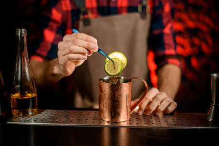 man bartender in red shirt and brown apron decorated metal cup by citrus slice using tweezers.の写真素材