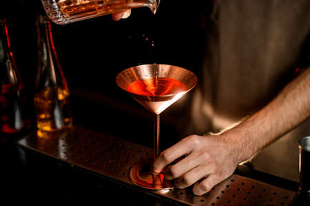 Close-up metal martini glass in which bartender in brown apron accurate pours bright cold alcoholic cocktail.の写真素材