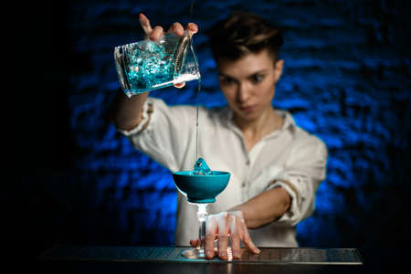 Woman bartender gently pours blue cocktail from mixing cup into glassの写真素材