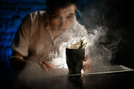 close-up of black ceramic old-fashioned glass with ice decorated with plants stands on smoky bar counter.の写真素材