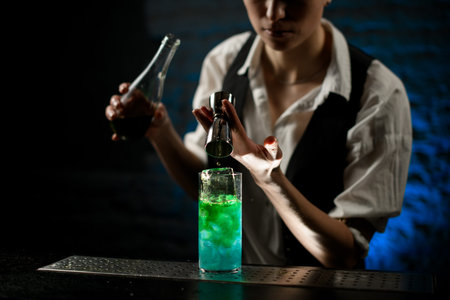 Young bartender pours drink for bright green cocktail from steel jigger into glass with ice.