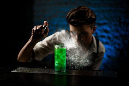 Young woman bartender sprinkles on illuminated glass with bright green cold cocktail and look at it.の写真素材