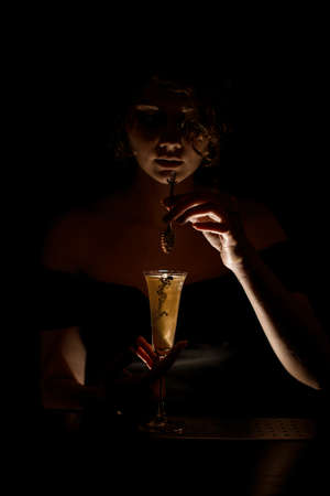 young attractive barman girl at dark bar holds spoon over glass with cocktailの写真素材