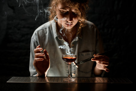 young woman bartender accurate holds tweezers with smoking wand near glass of cocktail.