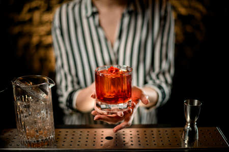 close-up old-fashioned glass with cocktail decorated with red pepper in woman hands at bar. Mixing cup and steel jigger stand nearby.の写真素材