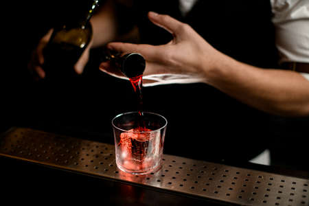 close-up. Barman's hand holds steel jigger and pours red liquid into glass with large ice cubeの写真素材