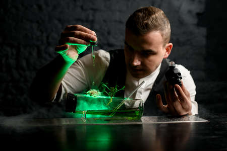 Bartender with dropper gently adds ingredient to special designer glass flask with smoky green canabis cocktail and decorated with plantsの写真素材