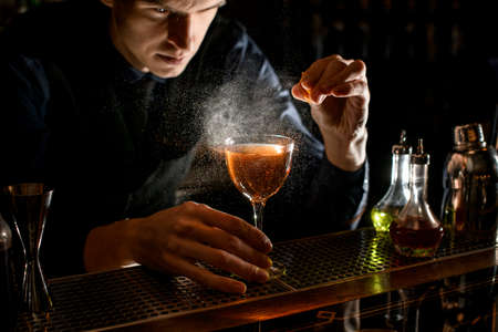 Young male bartender sprinkle to alcoholic cocktail with piece of citrus zestの写真素材