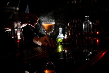 bartender's hand sprinkle to glass with amber alcoholic cocktail with piece of orange citrus at dark...