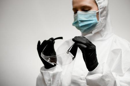 young woman in white protective overalls with hood holds safety glasses in her hands and looks at...