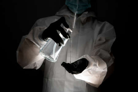 Person in protective clothing holds bottle of disinfectant and pours it on hand at dark roomの写真素材