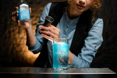 glass with cold blue drink stands at bar. woman bartender with jigger and bottle in hers hands...