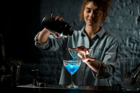 girl at bar pours bright blue drink from shaker into glassの写真素材